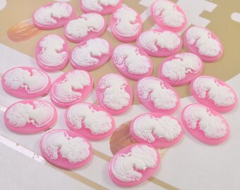 May include: A collection of pink and white cameo-style resin embellishments. The embellishments are oval-shaped and feature a white silhouette of a woman's profile.