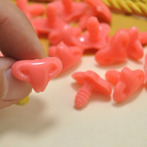 May include: A close-up of pink plastic safety eyes with a screw-on back. The eyes are in various sizes and shapes, some with a more rounded shape and others with a more pointed shape.