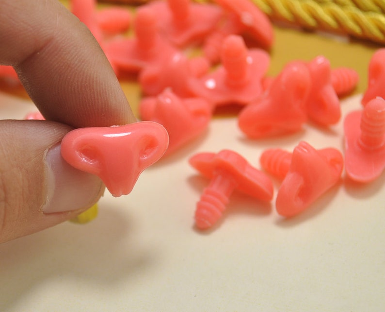 May include: A close-up of pink plastic safety eyes with a screw-on back. The eyes are in various sizes and shapes, some with a more rounded shape and others with a more pointed shape.