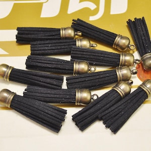 May include: A collection of ten black suede tassels with antique gold metal caps. The tassels are approximately 2 inches long and have a fringe-like appearance.