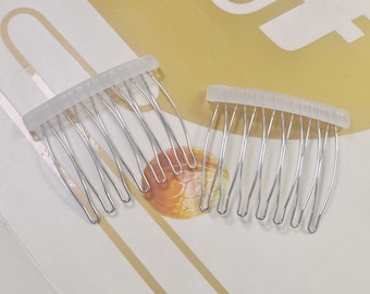 May include: Two silver-toned metal hair combs with clear plastic bases. The combs have curved rows of teeth, designed to secure hair. The image shows two combs, one slightly smaller than the other, likely for different hair styles or uses.