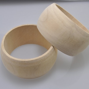 May include: Two unfinished wooden bangles. The bangles are light beige in color and have a smooth, rounded shape. The wood grain is visible on the surface. One bangle is lying flat, while the other is resting on top.