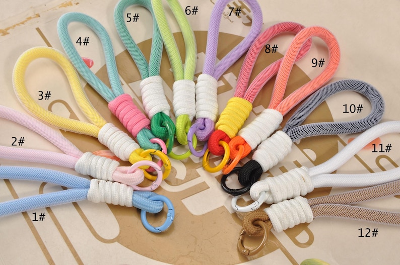 May include: A collection of colorful wrist straps in various shades, including blue, pink, yellow, green, and purple. Each strap features a woven cord loop and a matching or contrasting colored metal ring. The straps are numbered 1 through 12.