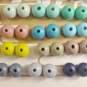 May include: Assortment of wooden beads in various colors, including brown, pink, yellow, blue, green, gray, and navy blue. The beads are arranged in rows on a white surface.