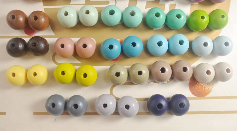 May include: Assortment of wooden beads in various colors, including brown, pink, yellow, blue, green, gray, and navy blue. The beads are arranged in rows on a white surface.