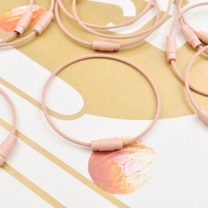 May include: A close-up of several pink metal keychains with a cable core. The keychains have a metal loop at one end and a metal cap at the other end.