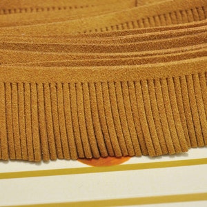 Pode incluir: Um close-up de uma guarnição de franja de camurça marrom. A franja tem aproximadamente 2,5 cm de comprimento e é feita de camurça macia e flexível.