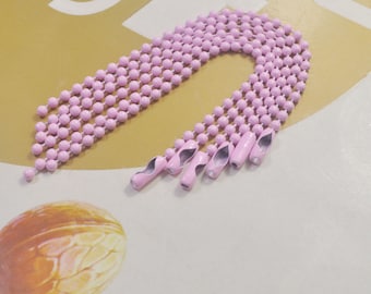 May include: A close-up of a pink metal chain with multiple loops. The chain is made of small, round beads and has a silver finish. The chain is lying on a light brown surface.