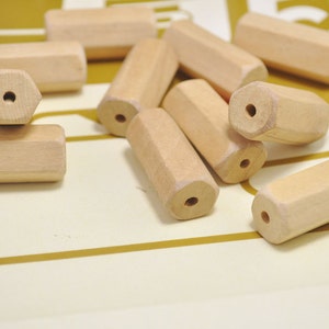 May include: A group of wooden beads in a natural color. The beads are hexagonal in shape and have a hole in the center. The beads are scattered on a light brown surface.