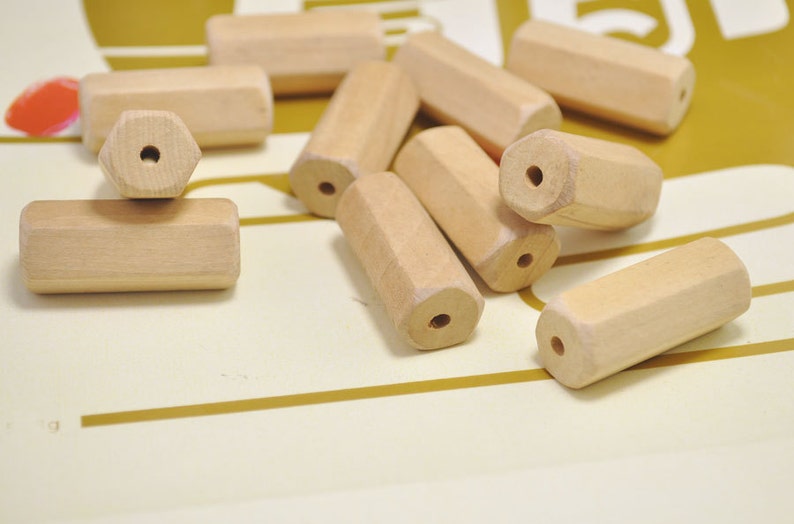 May include: A group of wooden beads in a natural color. The beads are hexagonal in shape and have a hole in the center. The beads are scattered on a light brown surface.