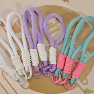 May include: A collection of colorful wrist straps in white, lavender, and teal. Each strap features a looped design with a wrapped base and a metal ring. The straps are arranged on a patterned surface.