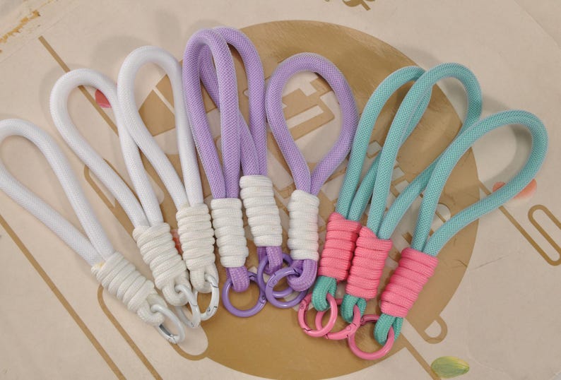 May include: A collection of colorful wrist straps in white, lavender, and teal. Each strap features a looped design with a wrapped base and a metal ring. The straps are arranged on a patterned surface.