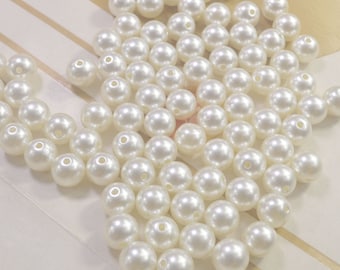 May include: A large quantity of white pearl beads, scattered on a white surface. The beads are round and smooth, with a slight iridescent sheen.