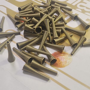 Könnte beinhalten: Ein großer Haufen goldfarbener Metallkegel. Die Kegel sind alle gleich groß und gleich geformt und in einem zufälligen Muster angeordnet. Die Kegel bestehen aus einem gewickelten Metalldraht.