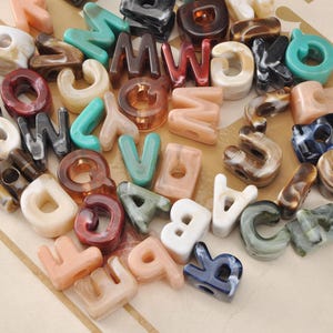 Op de afbeelding: Een verzameling geassorteerde letterkralen gemaakt van gemarmerde acrylhars. De kralen hebben verschillende kleuren, waaronder blauwgroen, bruin, wit, zwart en perzik, elk met een gladde, glanzende afwerking en een centraal gat om te rijgen.