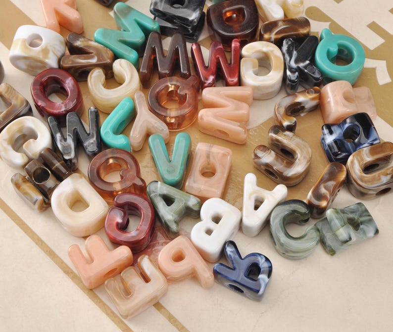 May include: A collection of alphabet letter beads in various colors and patterns. The beads are made of a translucent material, with colors including white, pink, brown, green, and blue. Each bead has a hole for stringing.