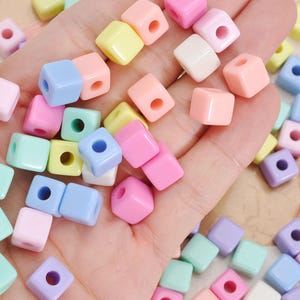May include: A collection of small, smooth, cube-shaped beads in a variety of pastel colors including pink, blue, yellow, green, and white. Each bead features a single centered hole, and the assortment is displayed in a scattered, loose arrangement.