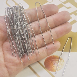 Op de afbeelding: Een close-up van een hand die een bundel zilveren haarspeldjes vasthoudt. De speldjes zijn golvend en hebben een puntig uiteinde.
