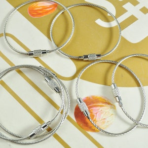 May include: Five silver cable key rings with a silver swivel clasp. The key rings are on a light brown surface.