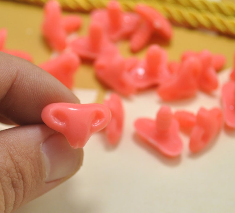 May include: A close-up of a pink plastic nose, with a small hole on each side. The nose is being held by a hand, and there are other pink noses in the background.
