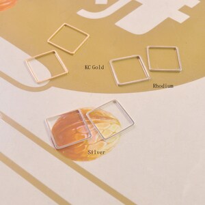 May include: Three square-shaped metal jump rings in different finishes: gold, rhodium, and silver. The rings are arranged in a row on a light brown surface.