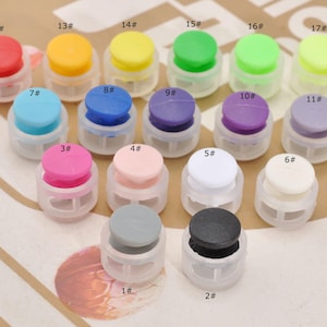 May include: A collection of colorful cord locks in various shades, including red, yellow, green, blue, and purple. Each cord lock has a round top and a clear, cage-like base. The image includes numbers next to each cord lock.