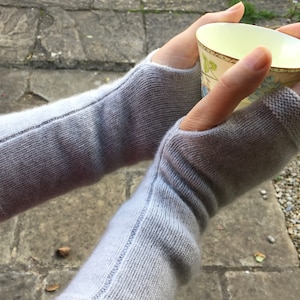 Könnte beinhalten: Hellgraue Fingerhandschuhe mit geripptem Bündchen und Nahtdetail. Die Handschuhe sind aus weichem Strickmaterial gefertigt. Die Person hält eine Teetasse mit floralem Design.