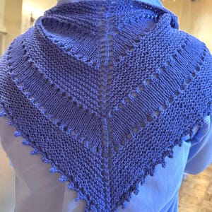 May include: A periwinkle-colored knitted shawl with a triangular shape. The shawl features a detailed openwork design and a scalloped edge. The knitted texture is visible, showcasing the craftsmanship of the handmade item.