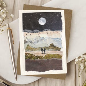May include: A rectangular greeting card featuring a collage of torn paper layers depicting a night landscape with a full moon, starry sky, mountain ranges, and a silhouette of two people holding hands, accompanied by a silver metal pen.