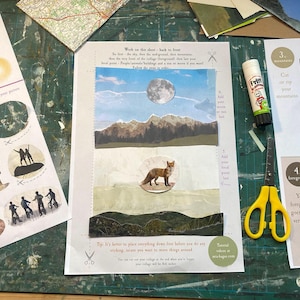 May include: A collage in progress featuring a landscape scene with a fox, mountains, and a full moon. The scene is created with cut paper and stickers. A tube of glue and yellow scissors are also on the green surface.
