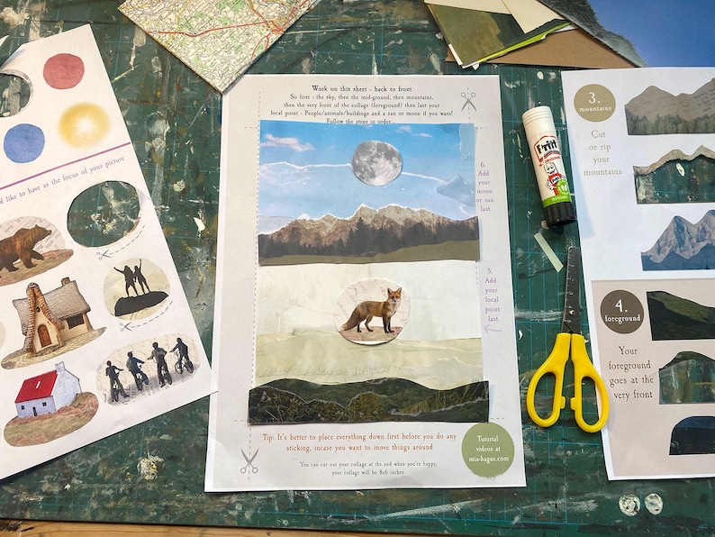 May include: A collage in progress featuring a landscape scene with a fox, mountains, and a full moon. The scene is created with cut paper and stickers. A tube of glue and yellow scissors are also on the green surface.