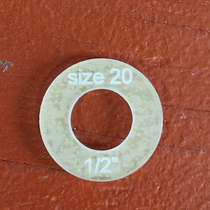 May include: A beige, circular washer with a central hole, resting on a reddish-brown surface. The text "size 20" and "1/2" are printed on the washer. The washer appears to be made of a durable material.
