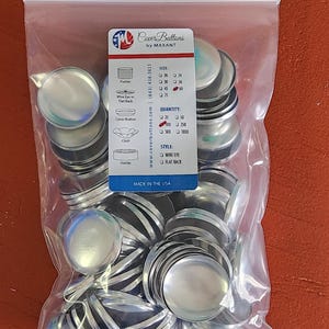May include: A clear plastic bag filled with silver metal cover buttons. A white and blue card with product details is visible. The card includes size, quantity, and style options. The text "Cover Buttons by MAXANT" is printed on the card. Made in the USA.