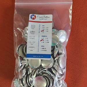 May include: A clear plastic bag filled with silver-colored cover buttons. The bag contains a label with the text "Cover Buttons by MAXANT" and details about sizes, quantities, and styles. The label also states "MADE IN THE USA".