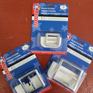 May include: Three packages of silver-colored belt buckles on a red wooden surface. Each package features a rectangular buckle with a pin, and the packaging includes the text "BUCKLES TO COVER" in English, French, and Spanish.