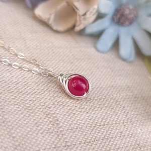 May include: A silver necklace featuring a small, round, red gemstone pendant. The gemstone is held in place by a wire setting. The necklace is presented on a textured, light-coloured surface, with blurred flowers in the background.