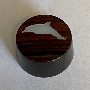 May include: A dark brown wooden knob with a light gray dolphin inlay. The knob has a cylindrical shape with a flat top and a slightly rounded edge. The dolphin is swimming to the right.