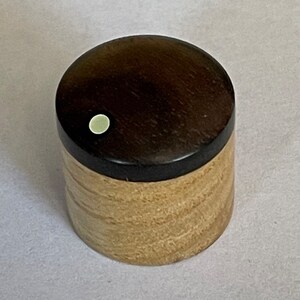 Könnte beinhalten: Eine kleine Holzkiste mit einem dunkelbraunen Deckel und einem hellbraunen Korpus. Der Deckel hat einen kleinen weißen Kreis in der Mitte.