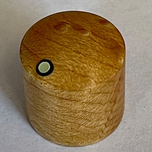 Puede incluir: Una pequeña caja redonda de madera con un acabado marrón claro. La caja tiene un círculo negro con un centro blanco en la parte superior.
