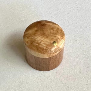 Puede incluir: Una pequeña caja redonda de madera con un patrón de veta de madera marrón claro. La tapa está hecha de una madera más clara con un patrón de remolino y tiene un pequeño acento dorado en el centro.