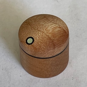 Puede incluir: Una pequeña caja de madera con una tapa redondeada. La tapa tiene un diseño de incrustaciones en blanco y negro. La caja está hecha de madera marrón claro.
