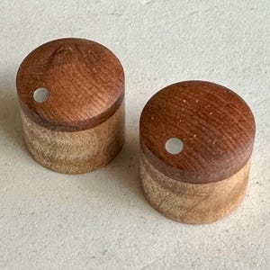 Puede incluir: Dos pomos de madera con una base de color marrón claro y una parte superior de color marrón oscuro. Cada pomo tiene un pequeño punto blanco en el centro.