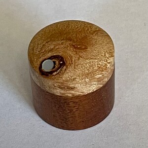 May include: A small, round wooden box with a light brown lid and a darker brown base. The lid has a white circle in the center.