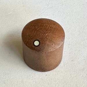May include: A cylindrical, wooden bottle stopper with a rounded top. The stopper is a medium brown color with a small, circular white accent on the top. The wood grain is visible on the surface.