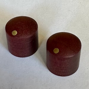May include: Two cylindrical, dark red wooden knobs with a small, round, gold-colored accent on the top. The knobs are smooth and have a slightly rounded top. The background is a plain, off-white surface.