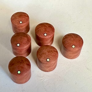 Puede incluir: Seis pomos de madera con un acabado marrón claro y un punto blanco en el centro. Los pomos están dispuestos en semicírculo sobre una superficie blanca.