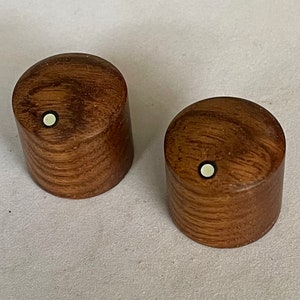 May include: Two cylindrical wooden knobs, displaying a rich brown grain. Each knob features a small, circular, off-white inlay on top. These knobs are likely for furniture or decorative purposes, with a diameter of approximately 3 cm.