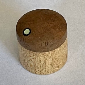 Puede incluir: Un pomo de madera con una base marrón claro y una parte superior redondeada marrón más oscura. La parte superior tiene un pequeño detalle circular blanquecino con un contorno negro. El pomo es probablemente para un mueble o un artículo similar.