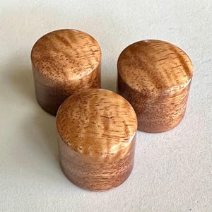 May include: Three wooden knobs with a natural wood grain pattern. The knobs are a light brown color with darker brown streaks.