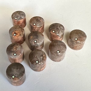 Puede incluir: Diez pomos de madera con un patrón de mármol marrón claro y gris y un punto blanco en el centro. Los pomos están dispuestos en semicírculo sobre una superficie blanca.